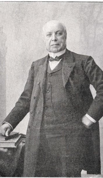An elderly Léon Harmel standing in a frock coat, one hand on his hip and the other resting on books; photograph published in the 1915 family memorial booklet.