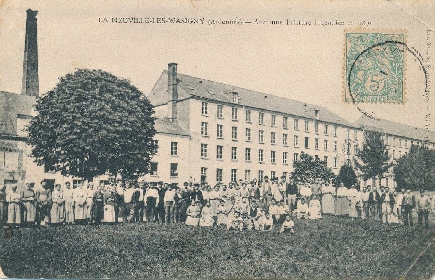 The large four-level building of the old spinning mill and its chimney; in front, on the lawn, a crowd of men, women and children poses for the photographer.