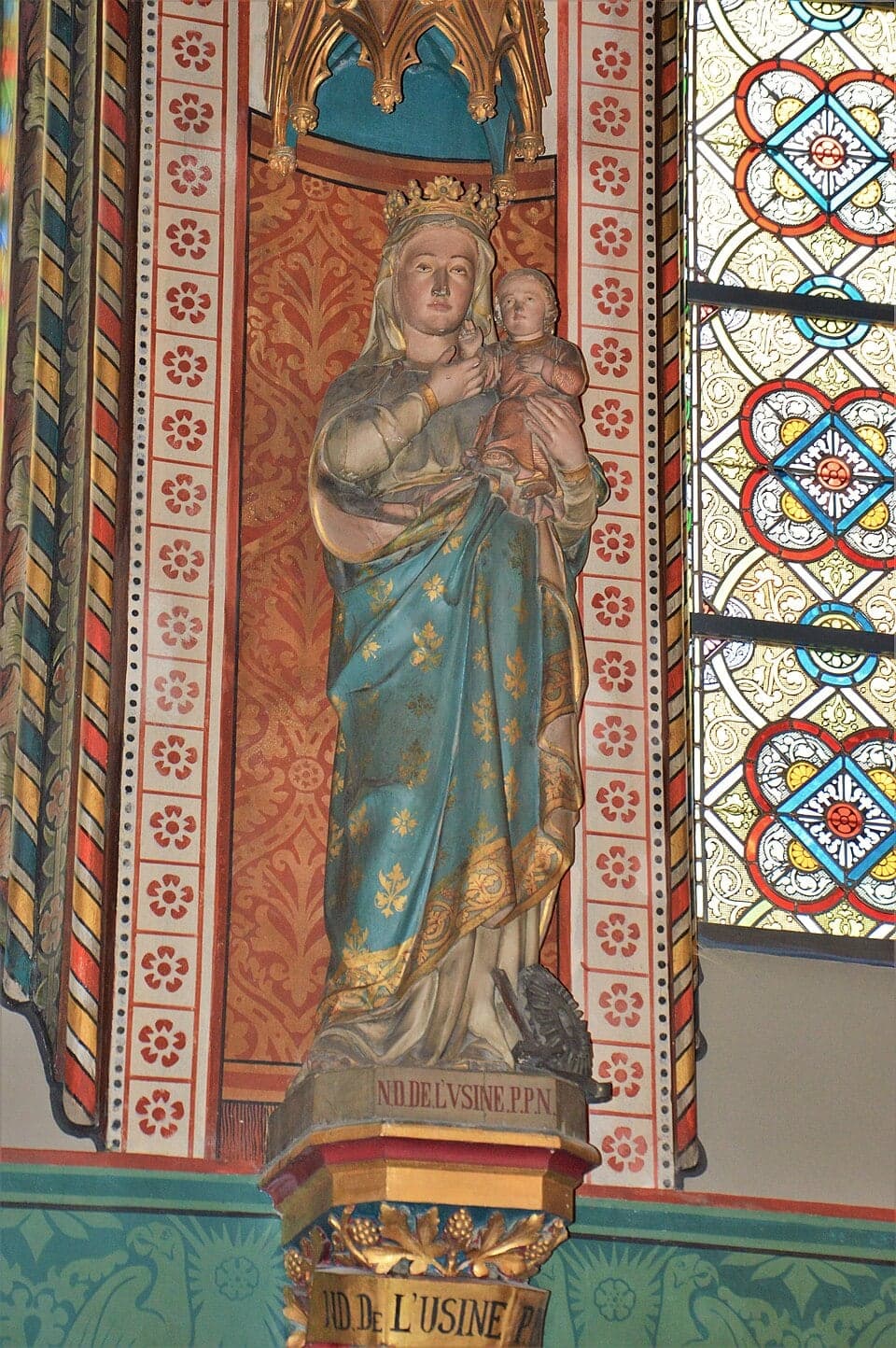 Statue of the crowned Virgin holding the Child, with a cogwheel at her feet; on the base, the inscription “N.D. de l'Usine”.