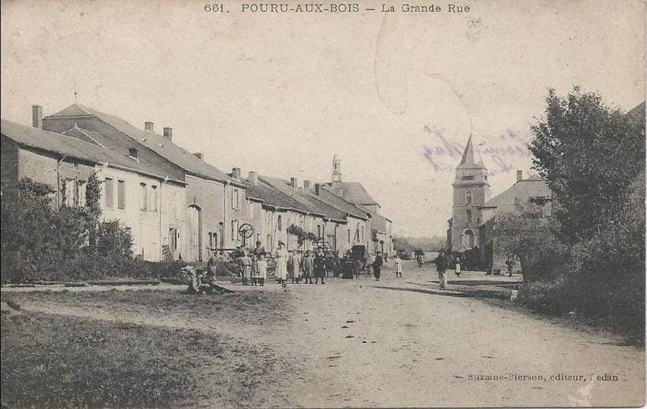 The high street of the Ardennes village, lined with low houses, with villagers gathered in front of the church with its pointed steeple.