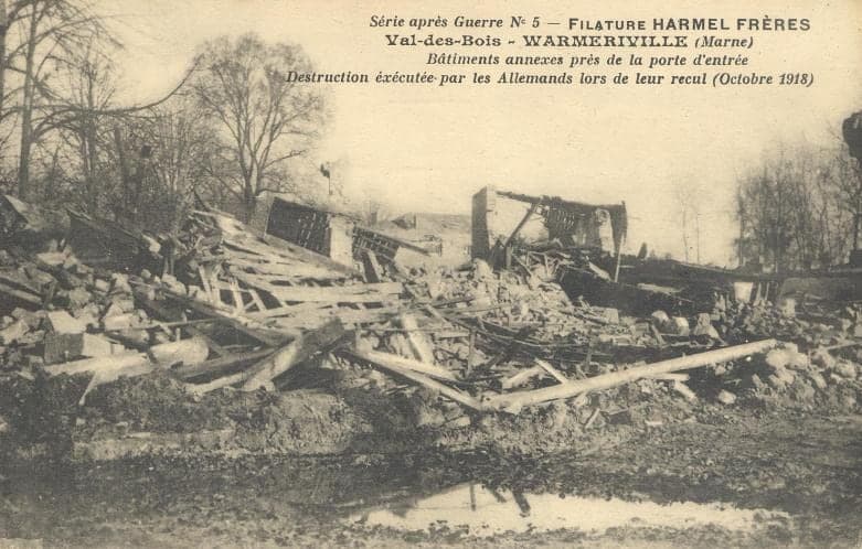 Collapsed roof frames and tangled planks of the outbuildings, beside a puddle, under bare trees.