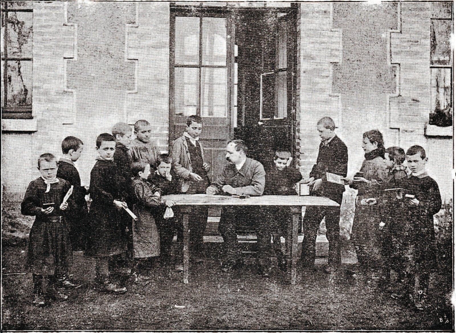 In front of a brick house, schoolboys in smocks crowd round a table where a teacher records their deposits; several hold their savings books.
