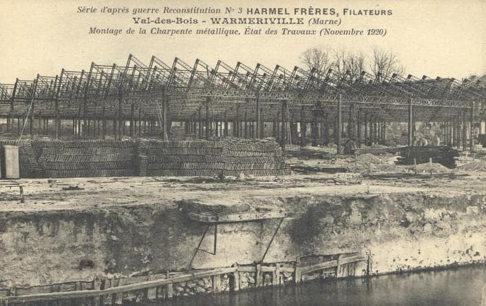 The new steel roof frame of the workshops rising over a vast building site, above piles of materials, by the water.
