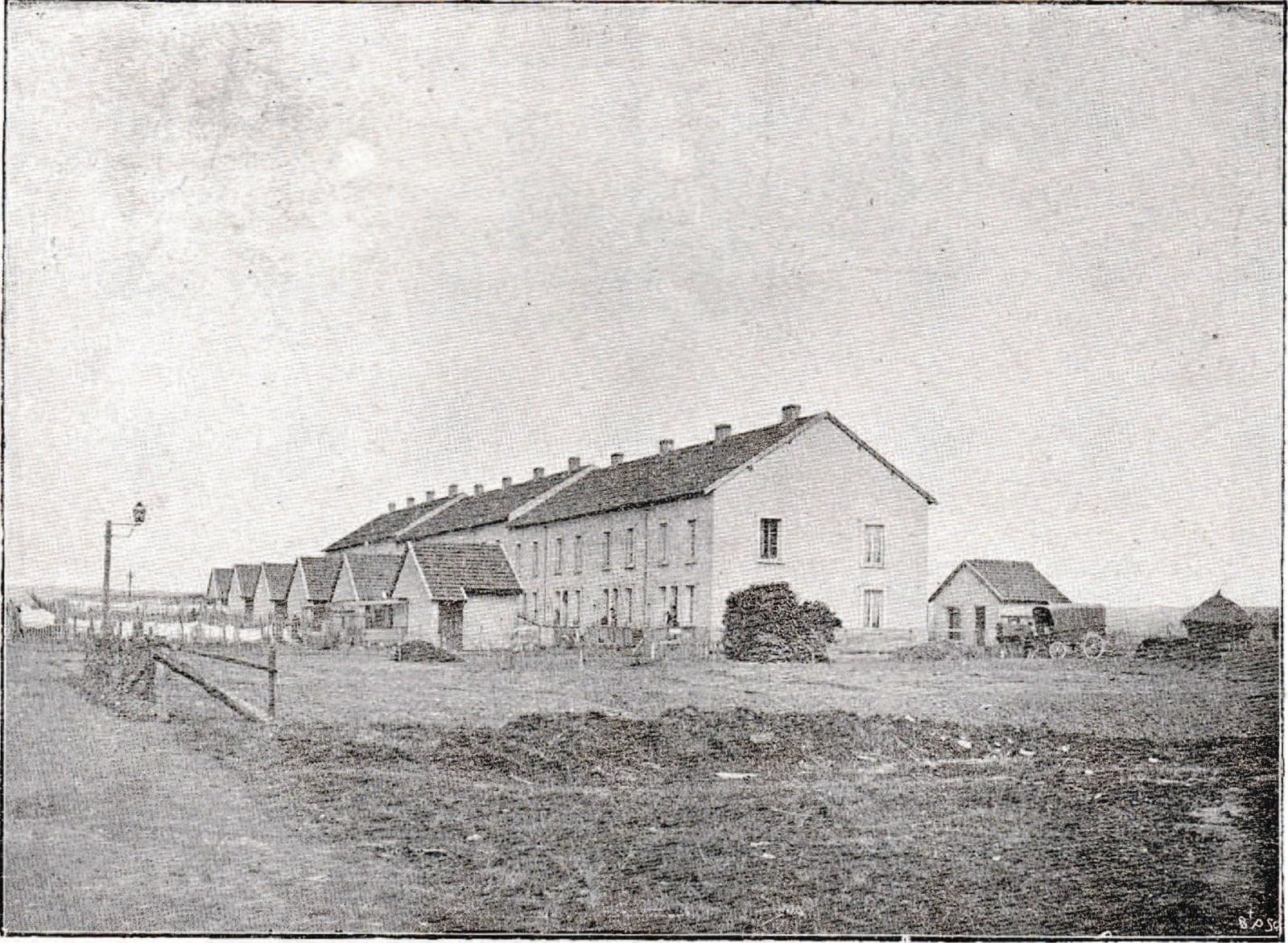 A long row of two-storey workers' houses, extended by lower houses, at the edge of still bare ground; a street lamp stands on the left.