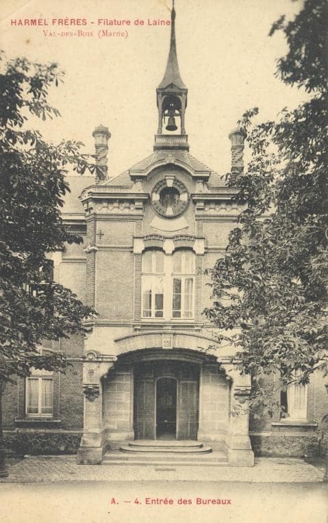 The brick façade of the mill offices, with its arched doorway, clock and bell turret, between two trees.