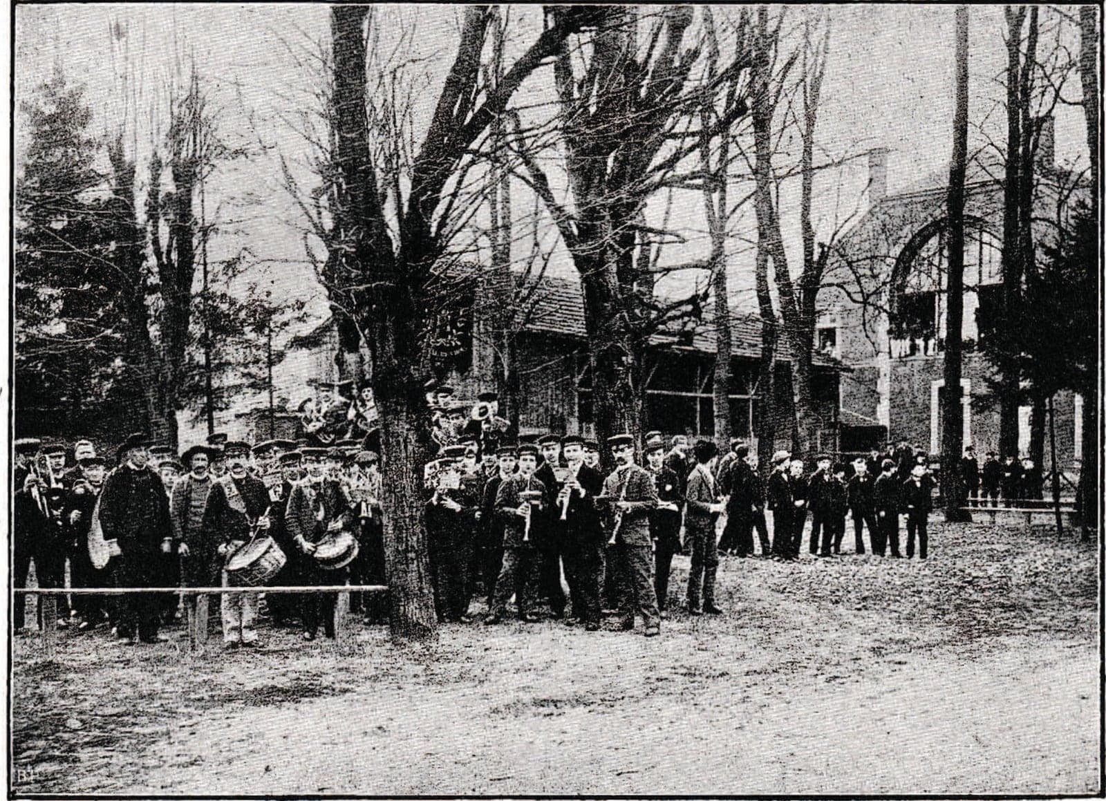 In the tree-lined courtyard, the band in caps, with drums and brass, stands before the workshops; children and workers look on from behind a rail.