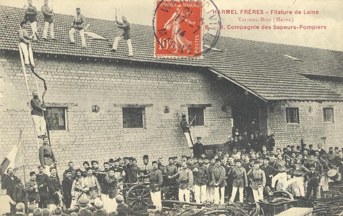 The mill fire brigade at drill: uniformed men climb ladders and stand on a workshop roof, while the company and the hand pump are gathered in front.