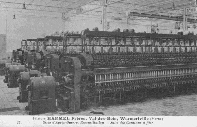 A row of continuous spinning frames, with their motors in the foreground, in a hall with a steel roof frame.