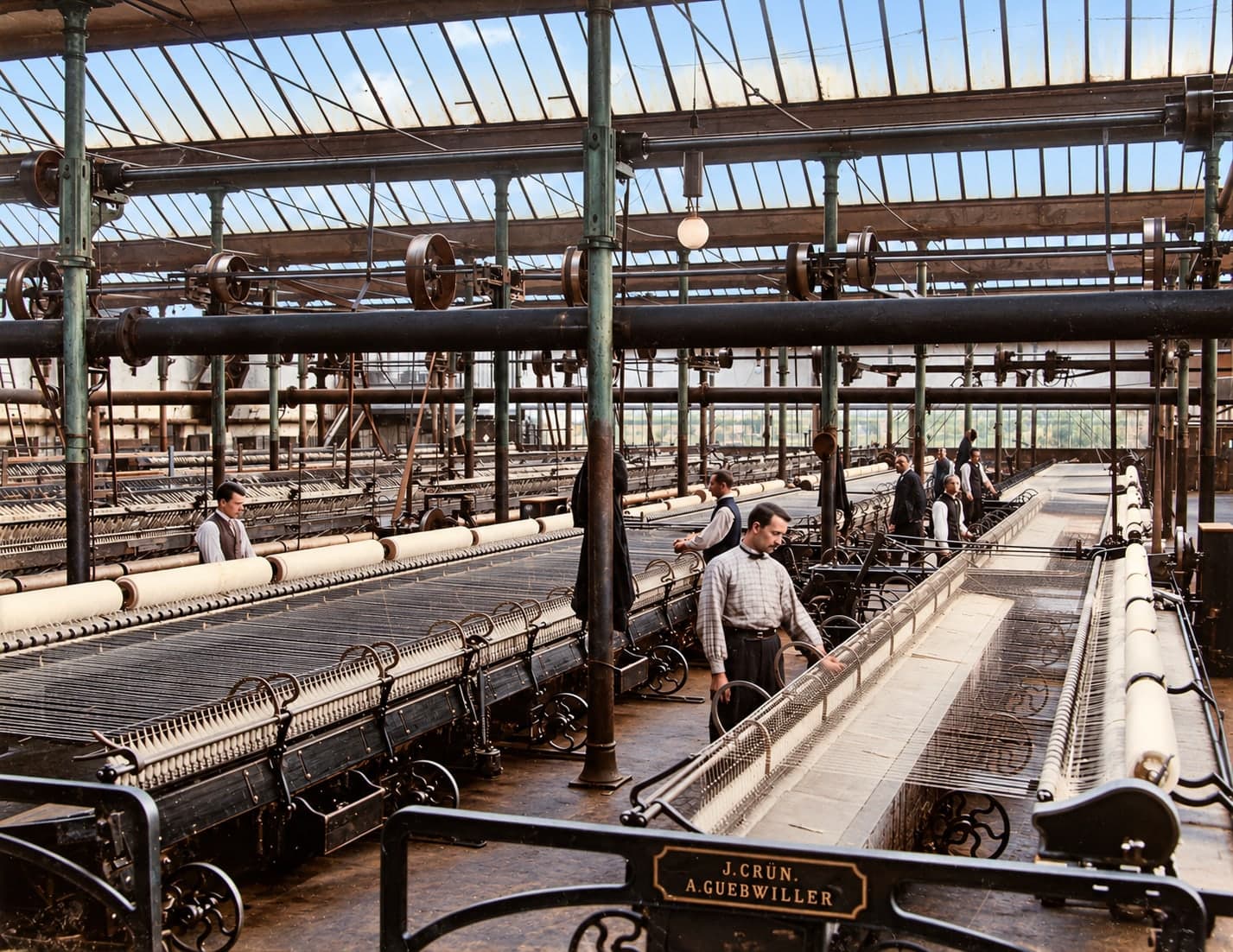 A long room under a glass roof: rows of spinning machines; a worker in an apron stands in the foreground, others are working in the background.