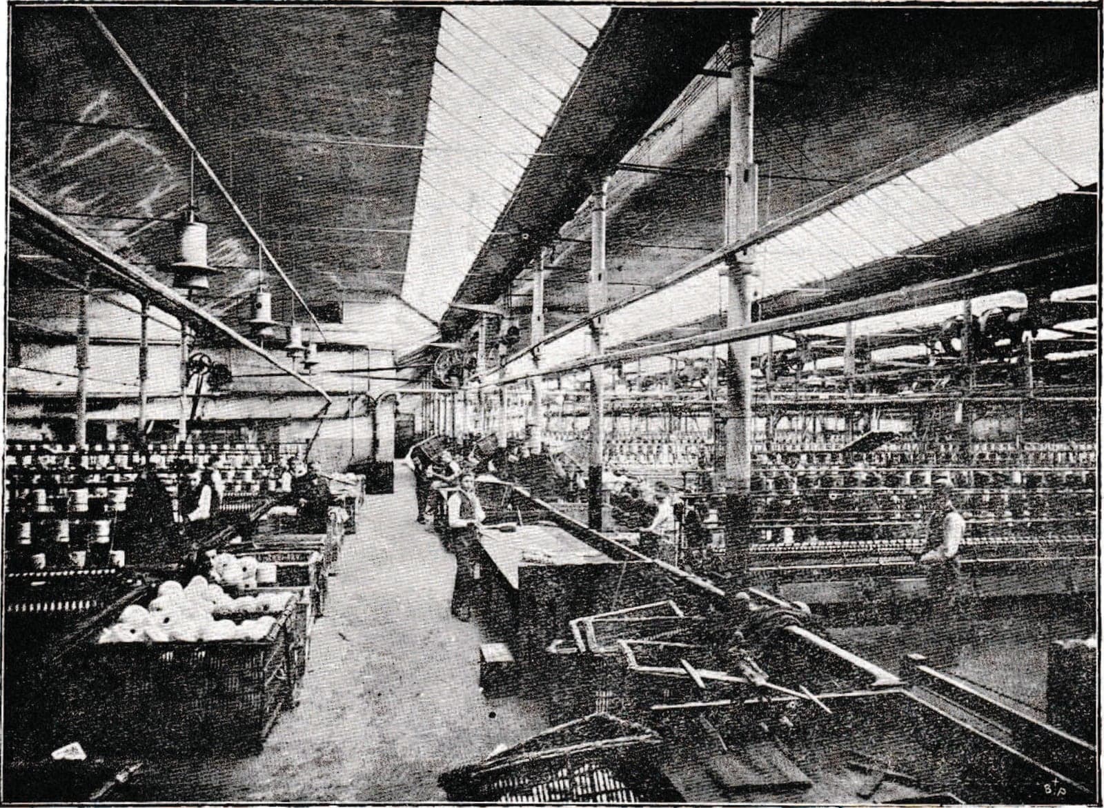 A long spinning hall under a glazed sawtooth roof: rows of frames, line shafts, crates of wool bobbins in the foreground and workers at their posts.