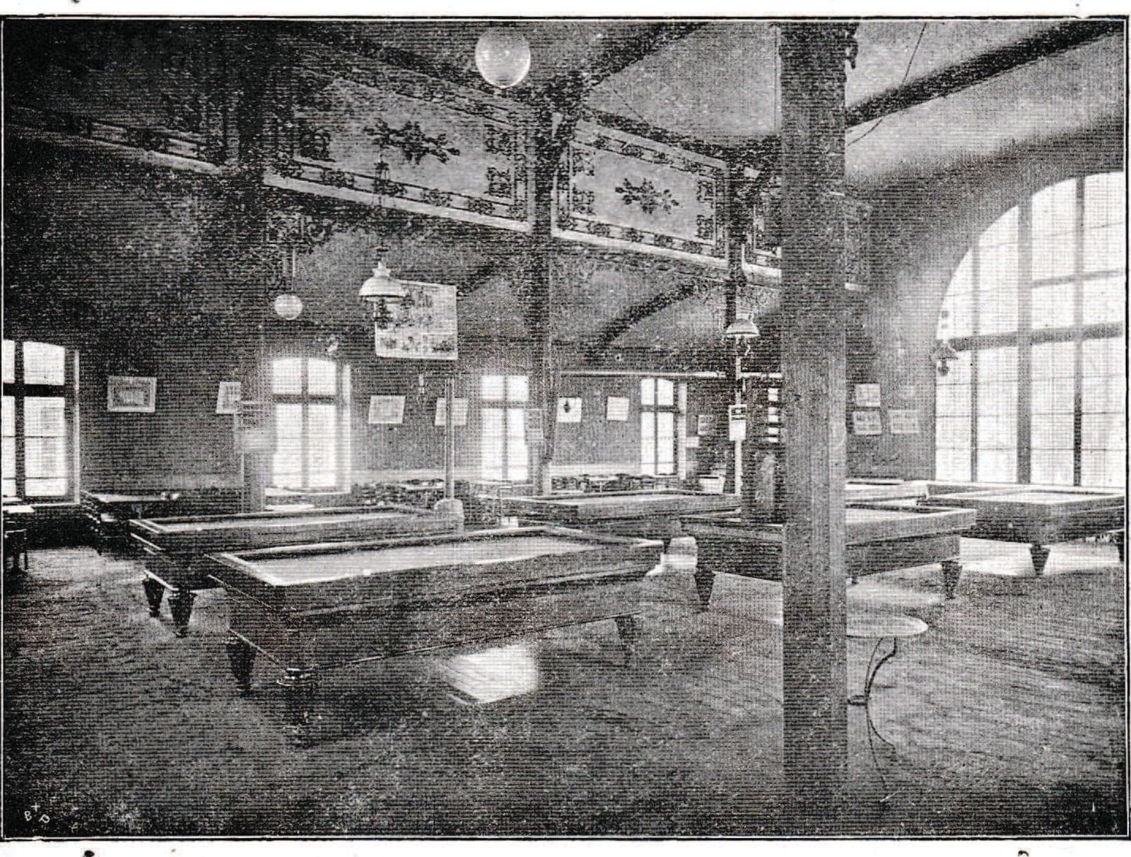 The large hall of the Circle, its walls decorated with friezes and lit by a tall arched window: several billiard tables in a row, hanging lamps and framed prints.