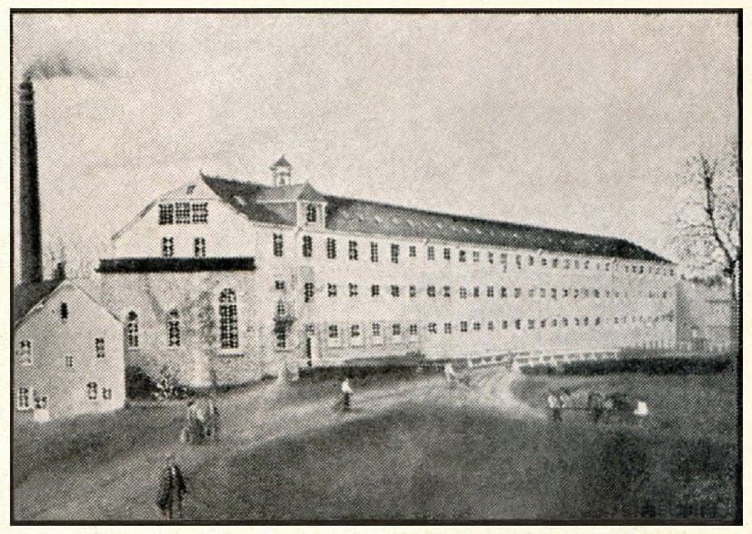The large four-level building of the old mill, with its central gable topped by a small bell turret, a chimney on the left and a few people walking on the path.