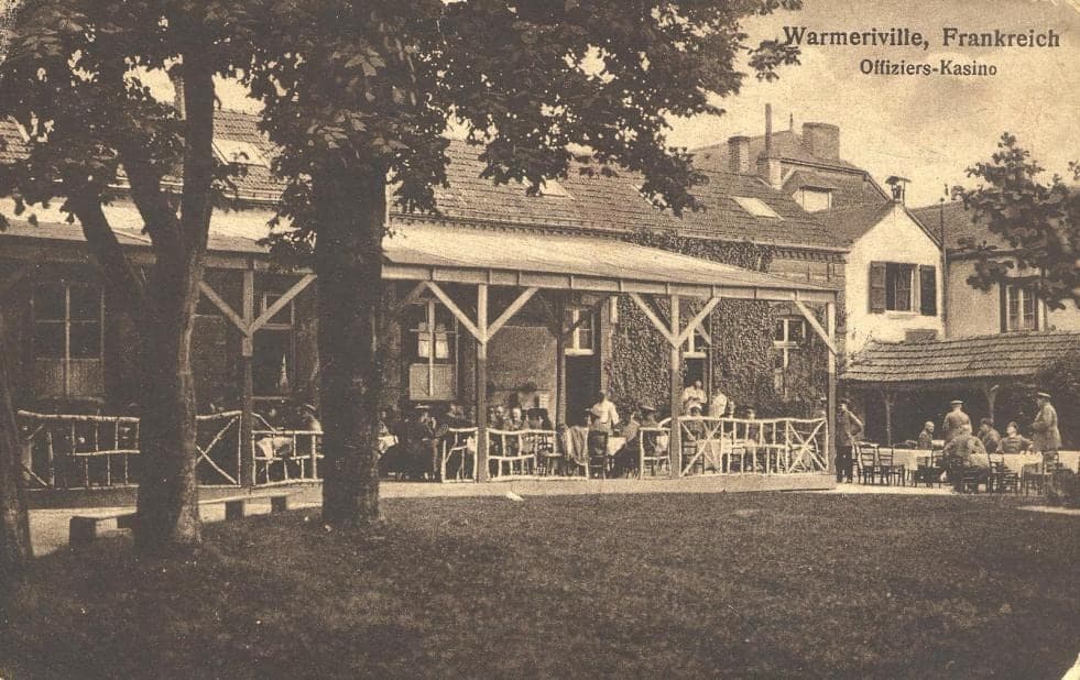 Under the trees, a covered terrace set with tables where German officers sit, in front of a house covered with Virginia creeper.
