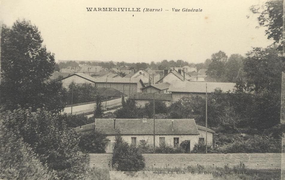 The roofs of Warmeriville seen from a wooded height, with a long low building and a road lined with posts in the foreground.